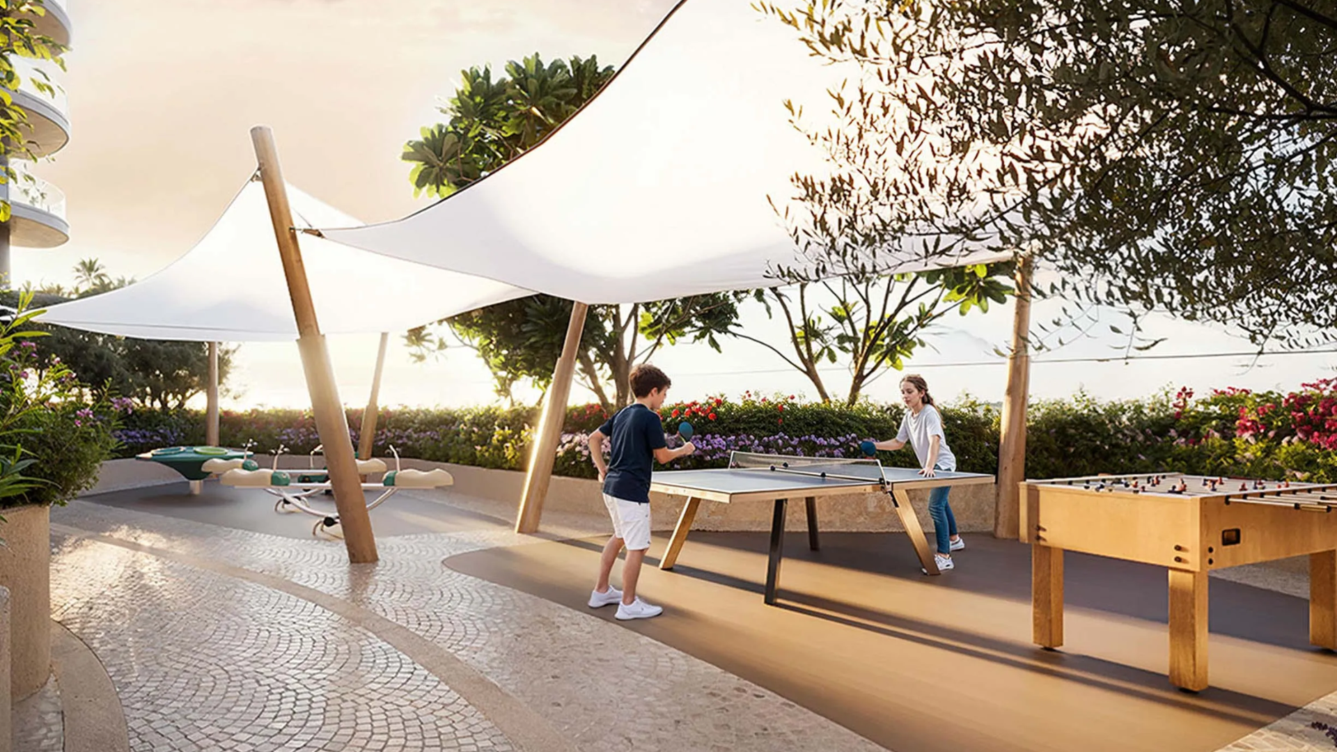 Children playing table tennis in a shaded outdoor recreation area.