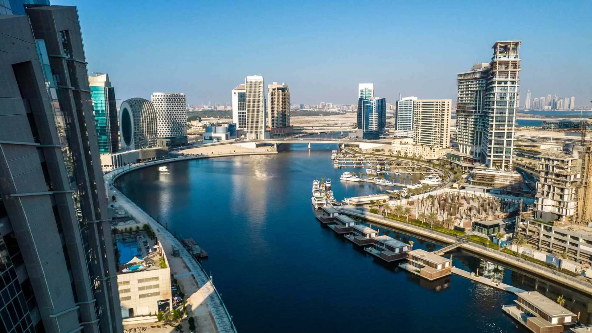 An urban waterfront with floating villas and yachts, framed by high-rise residential buildings under a blue sky.