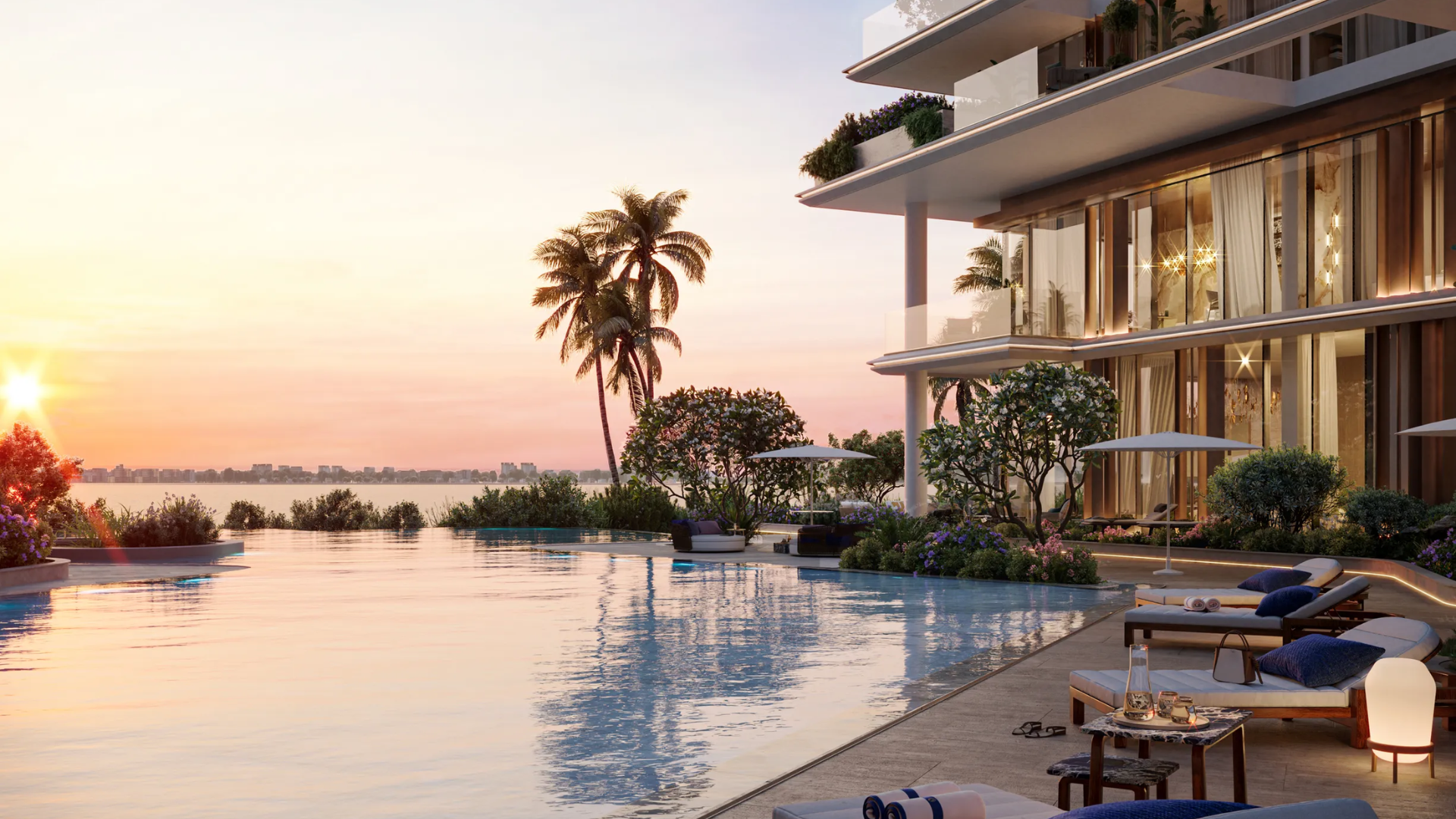 A resort-style swimming pool with lounge chairs and palm trees at sunset overlooking a large body of water.