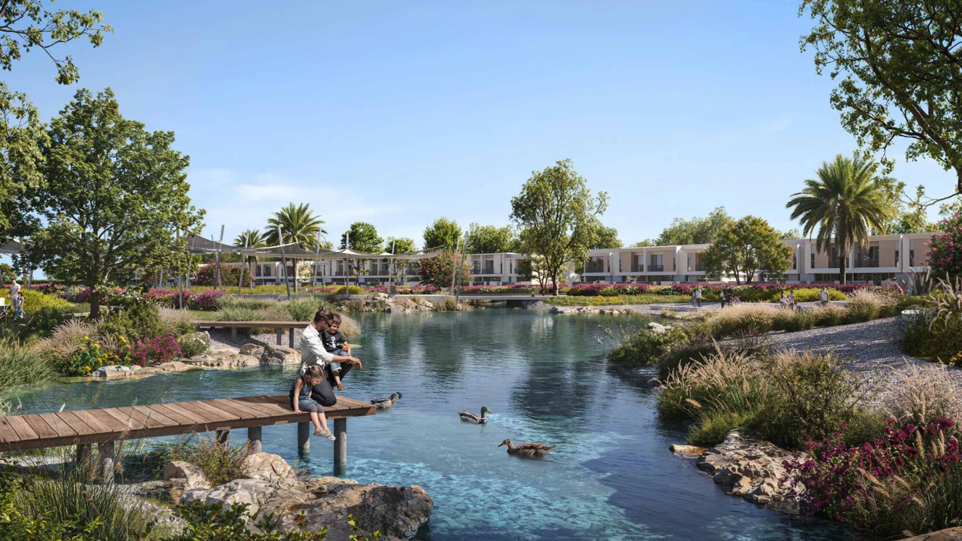 Residential park area featuring a clear water pond surrounded by flowering plants and townhouses in the background.