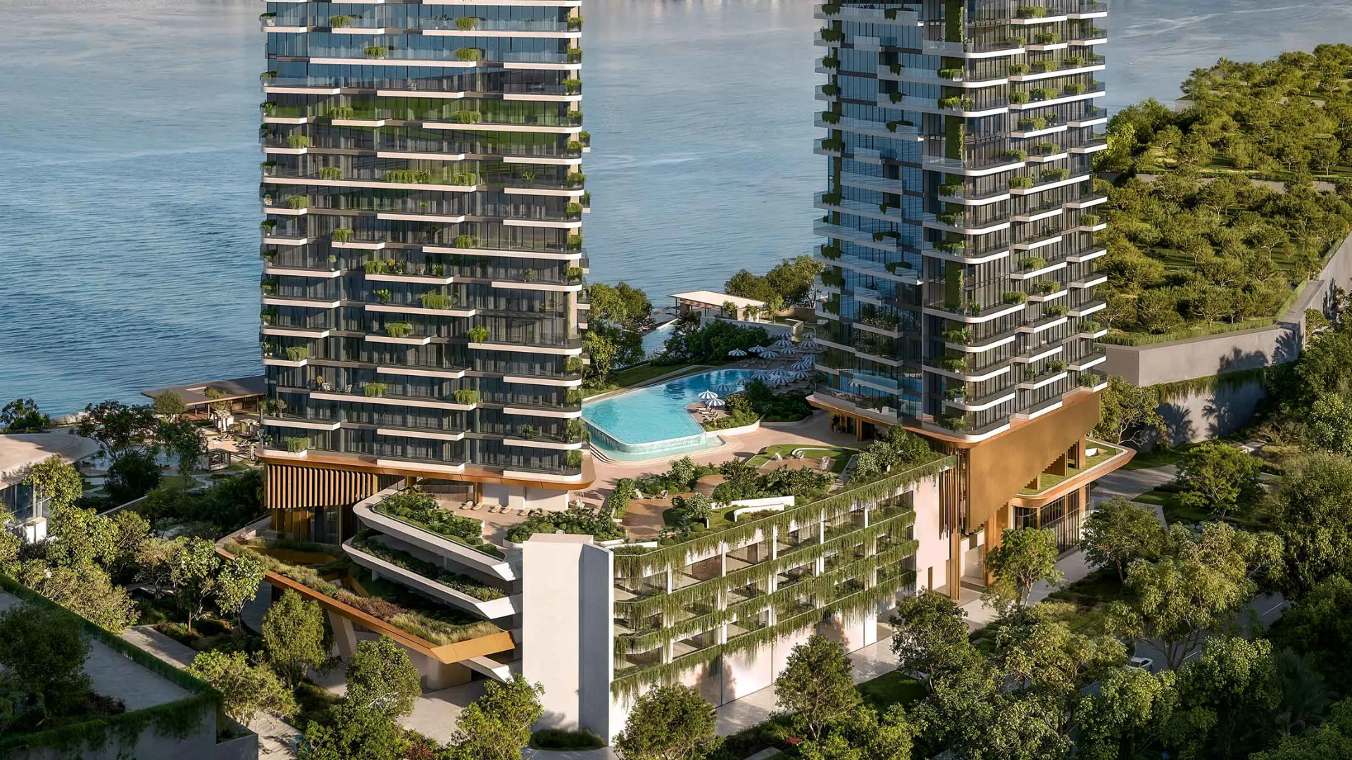 Two modern waterfront residential towers with lush green terraces and a large swimming pool deck overlooking the sea, surrounded by dense greenery.