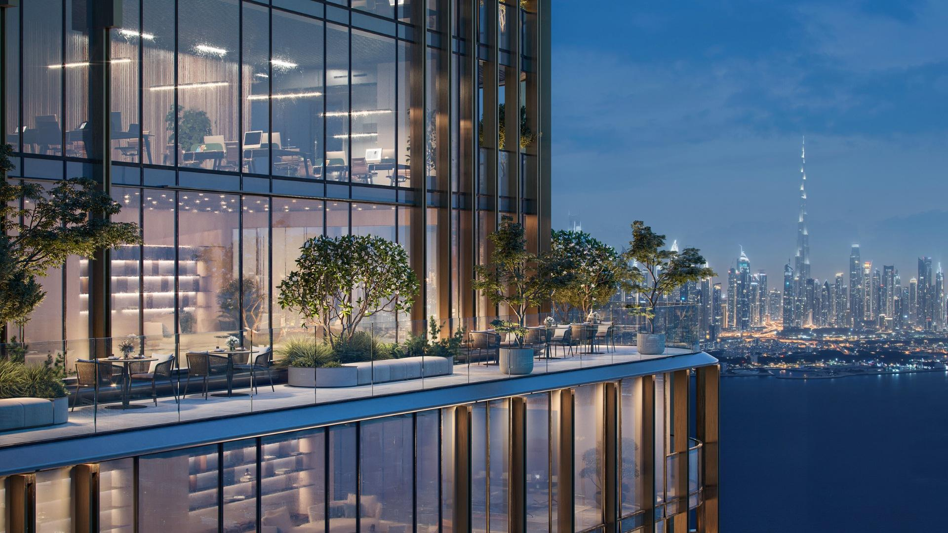 Glass-fronted building with a shared outdoor patio, captured during the evening with lights visible inside and across the water.